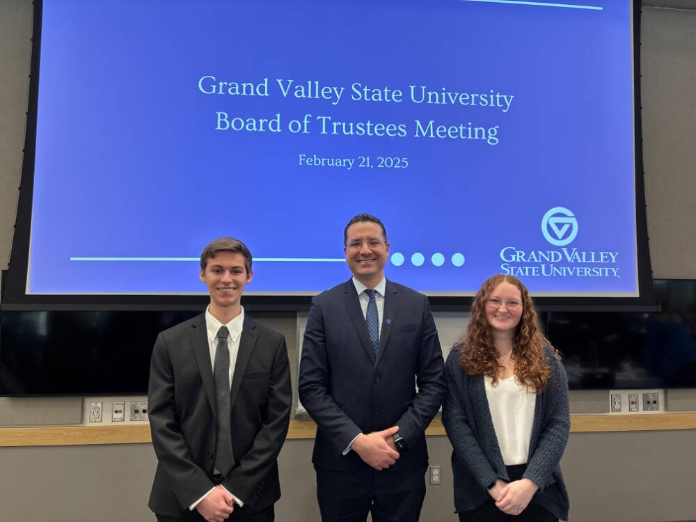 Dean Marouane Kessentini,in the middle, alongside students Natalie Kline,to the right and Marcos Sanson, on the left stand in front of a GVSU blue drop banner with "Grand Valley State university  Board of Trustees Meeting written on it.